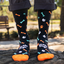 Load image into Gallery viewer, This image shows a person wearing black Guide Dogs socks decorated with orange, green and white paw prints and dog bones. The toes are orange, and "Guide Dogs" is written in white across the top of each foot.
